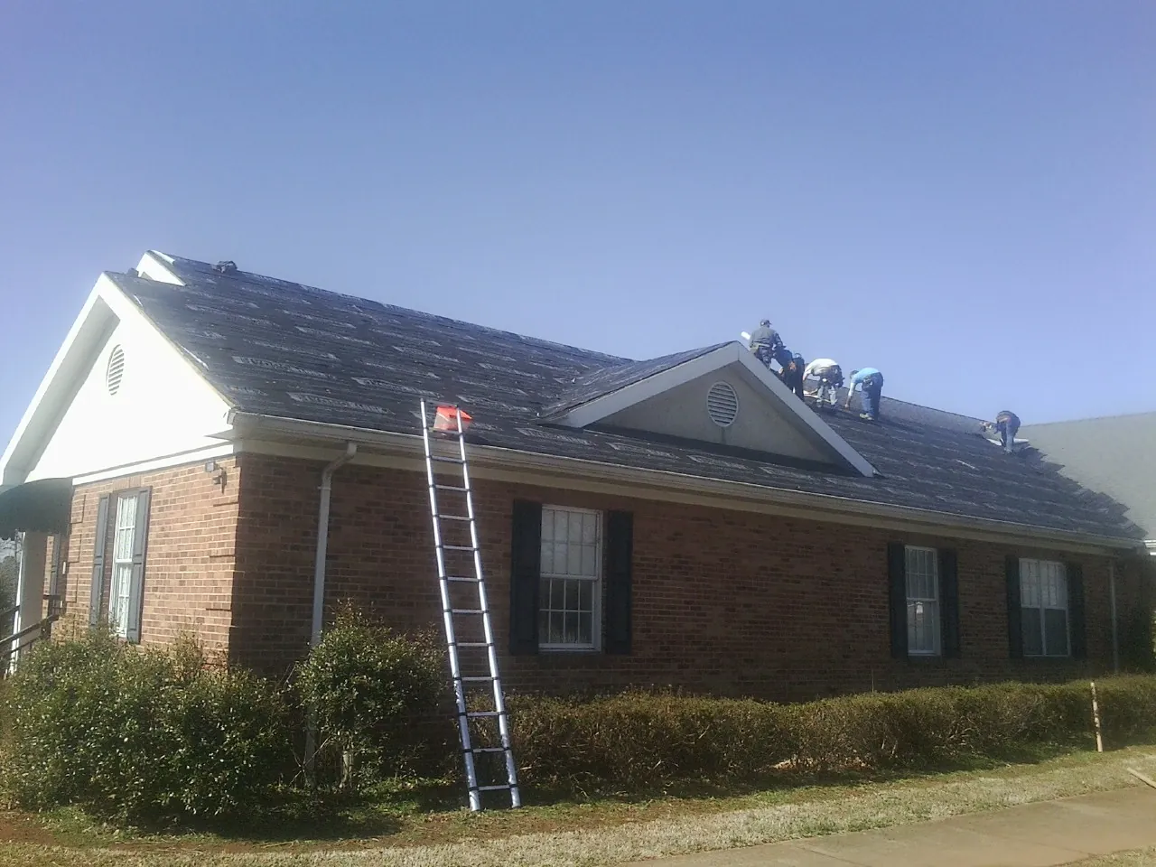 Crew tearing shingles off a brick ranch roof with a ladder set — Emergency Roof Repair work by our crew in Wonalancet