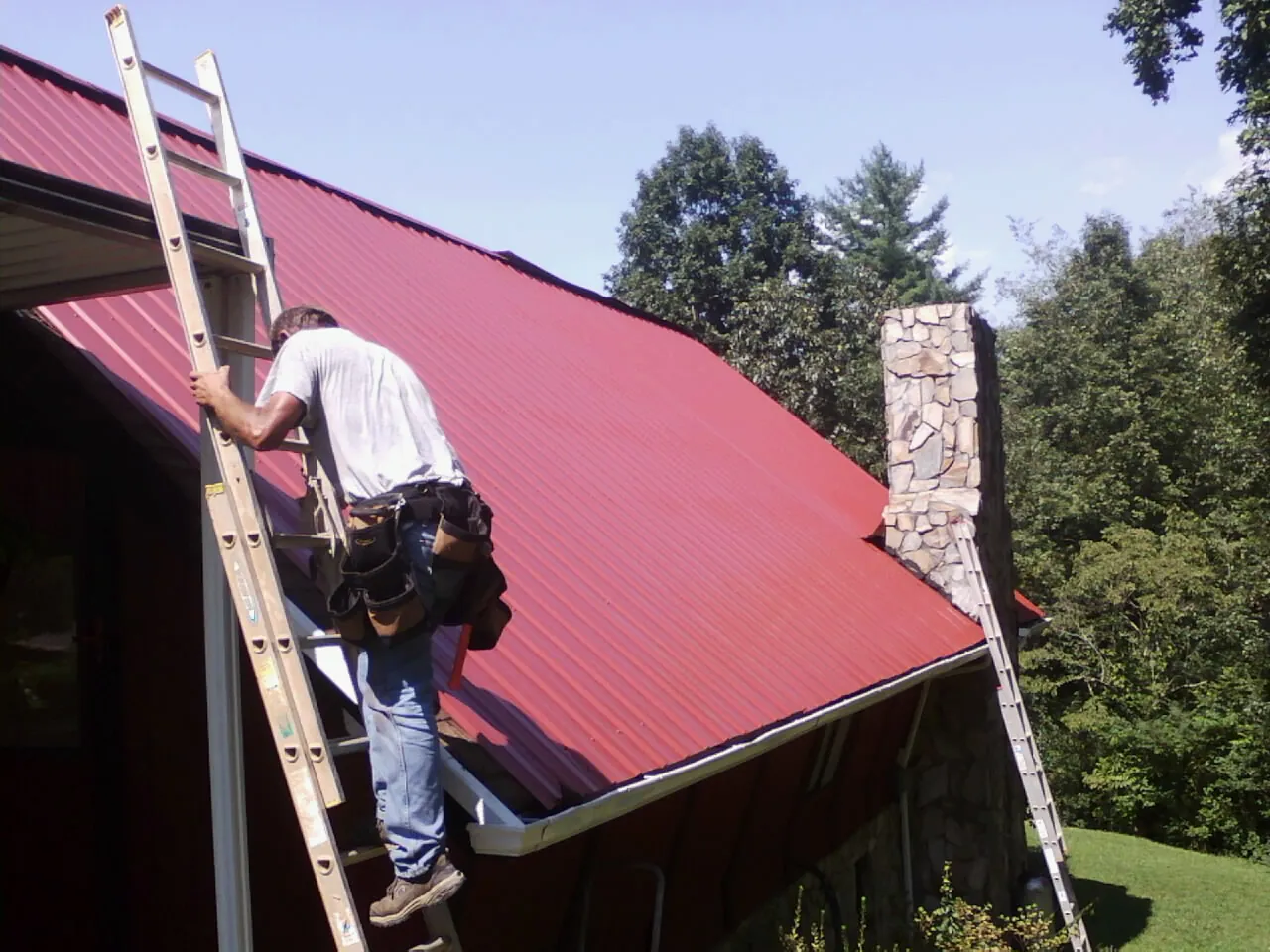 Roofer on a ladder beside a red metal roof and stone chimney — Soffit & Fascia Repair by Roofing Brothers in Wonalancet